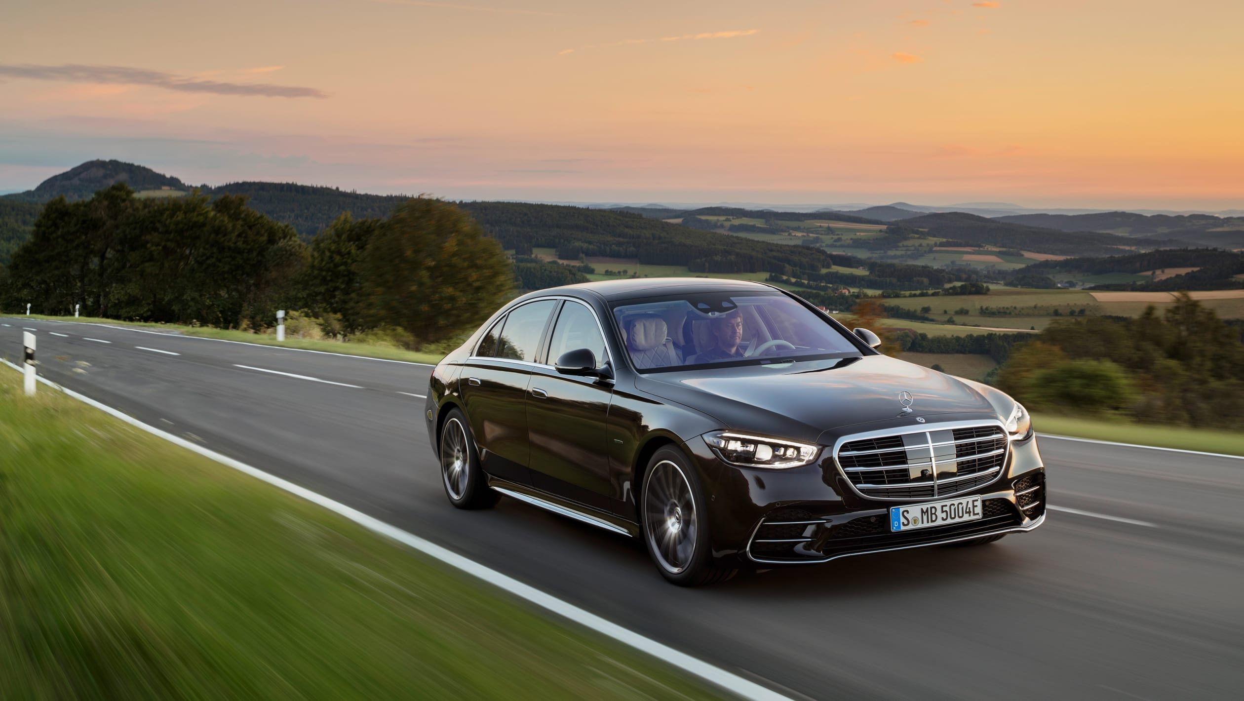 Black S Class car driving on a scenic road with natural landscape in the background. Our luxury chauffeur service provides comfortable and stylish transportation with breathtaking views.